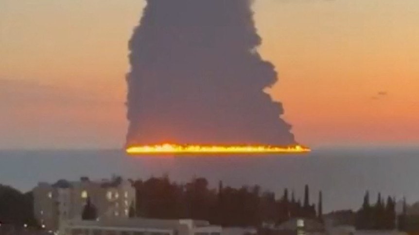 Flame line and smoke column over the water, houses and trees in the foreground.